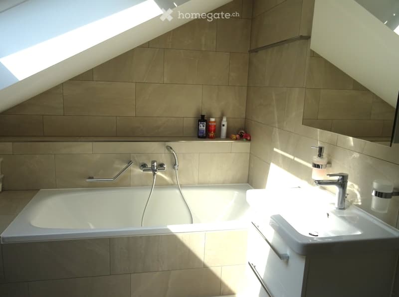 Modern bathroom with a bathtub, sink, and handrail, featuring beige tiles and natural light from a skylight.