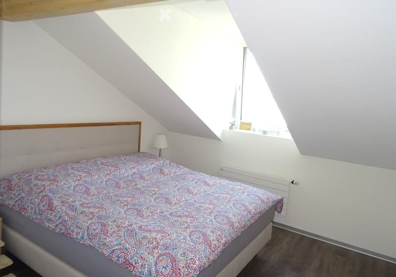 Cozy attic bedroom with a single bed, patterned bedspread, bedside lamp, and a small shelf with decorative items.