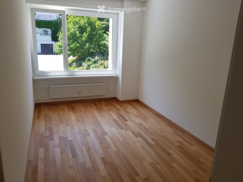 Empty room with wooden flooring, white walls, and a large window with a view of greenery outside. A radiator is visible below the window.