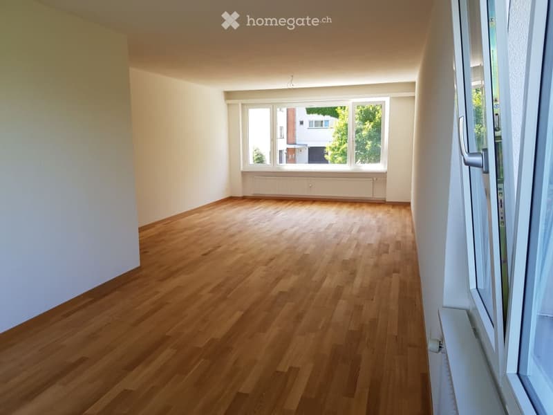 Empty room with wooden flooring, white walls, and large windows with a radiator below them.