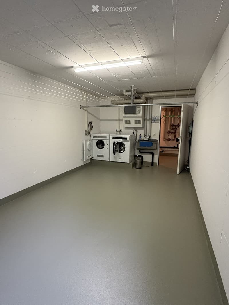 Laundry room with a washing machine, sink, and visible plumbing. The space is clean and functional with a concrete floor and white walls.