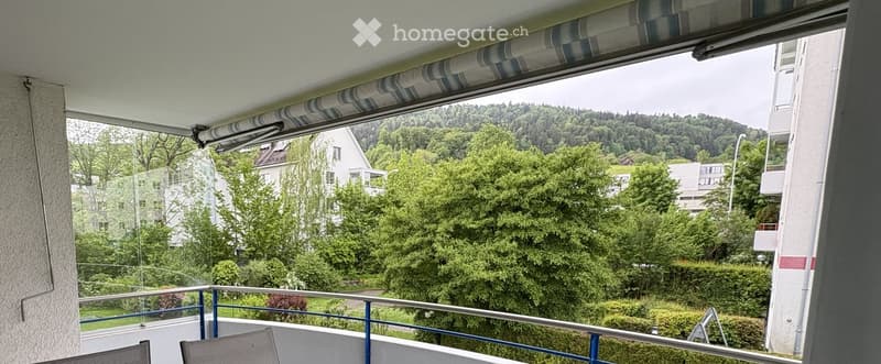 Balcony with metal railing, striped awning, and outdoor chairs overlooking a green, hilly landscape with trees and buildings.
