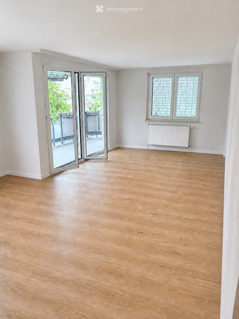 Empty room with light wooden flooring, white walls, large windows, and a sliding glass door leading to a balcony.