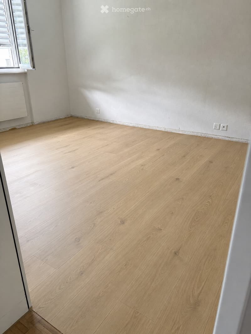 Empty room with light wooden flooring and white walls, featuring a window with blinds and a radiator on the left side.