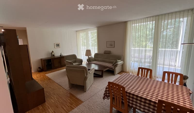 Bright living room with large windows, wooden flooring, a sofa, armchairs, coffee table, dining table with chairs, and a cabinet with a lamp and picture frame.