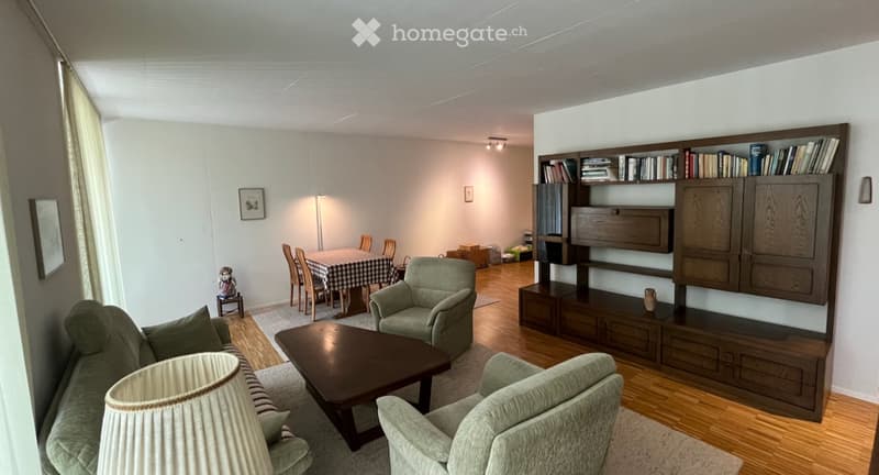 A well-furnished living room with a sofa, coffee table, armchairs, a bookshelf, and a dining area with a table and chairs.
