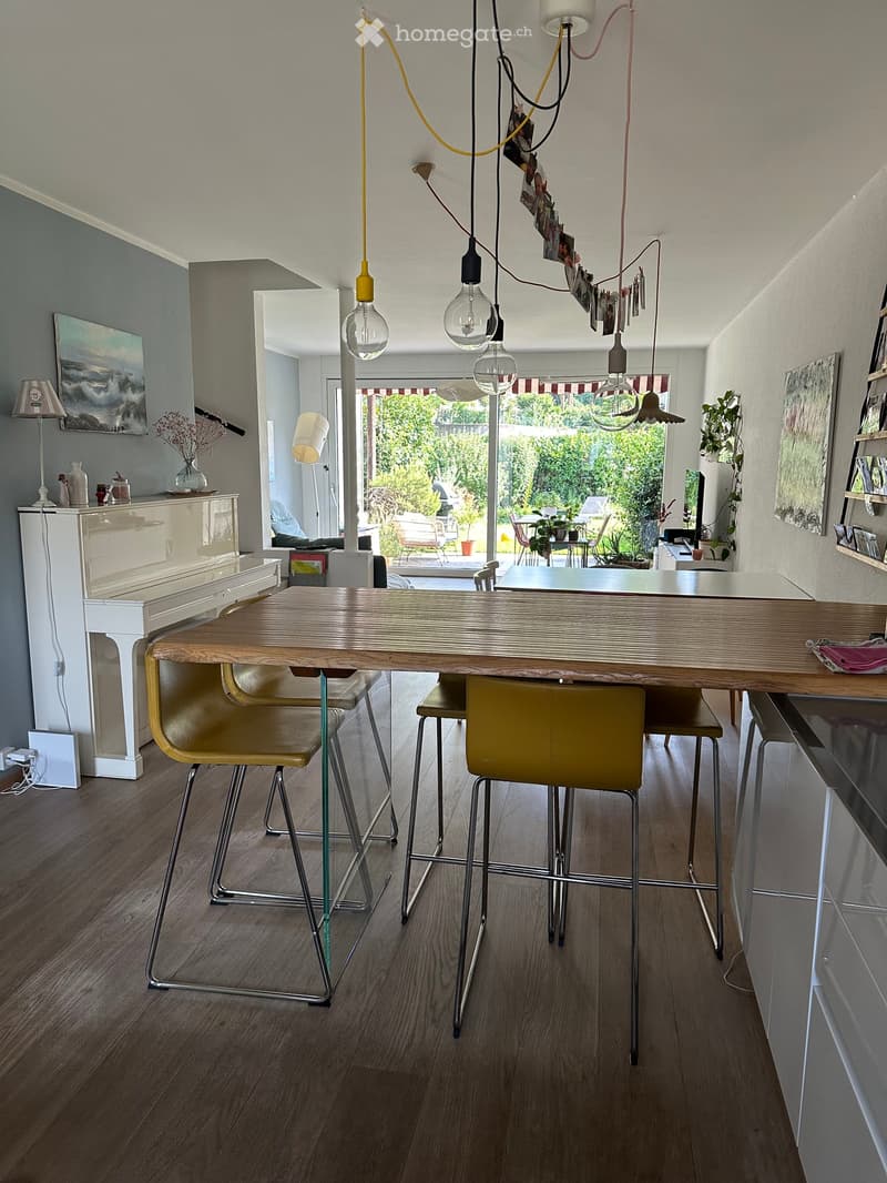 Modern kitchen with wooden floor, central island table with four yellow stools, pendant lights, white cabinetry, and a view to a garden through large windows.