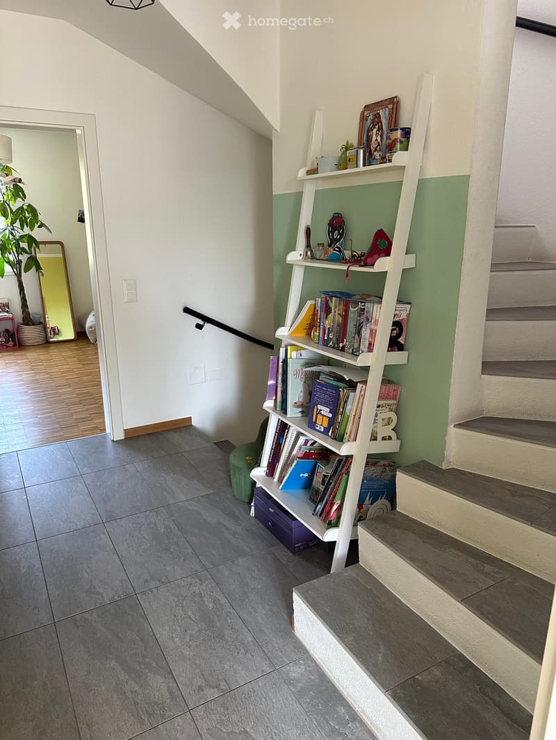 Hallway with gray tiled floor, white walls, staircase leading upwards, white bookshelf with books and decorative items, green accent wall, and a small green stool near the bookshelf.