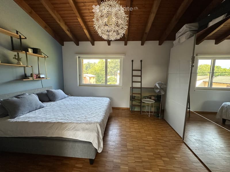 Cozy attic bedroom with wooden floors, slanted wooden ceiling, large windows, bed with pillows, shelves, fan, and ladder leading to loft area.
