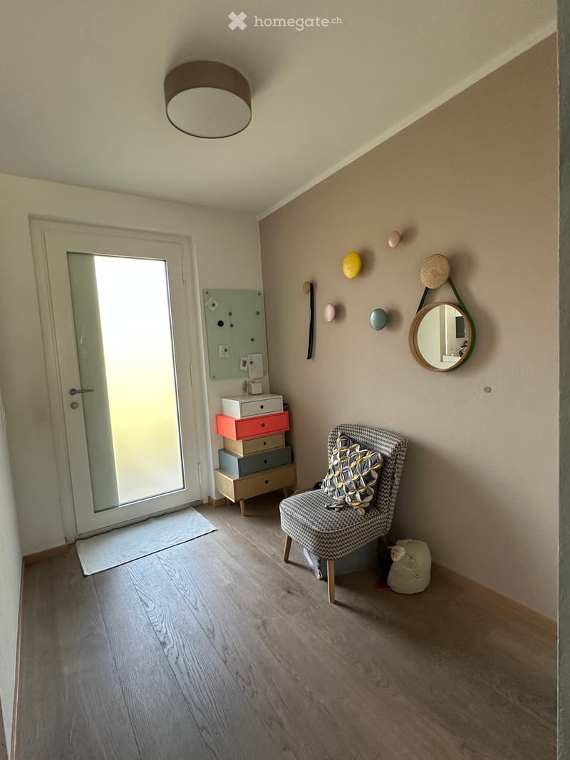 A small, modern hallway or entryway with light wooden flooring, a hanging light fixture, a patterned chair, a round wall mirror, and a stack of colorful storage boxes near the door.