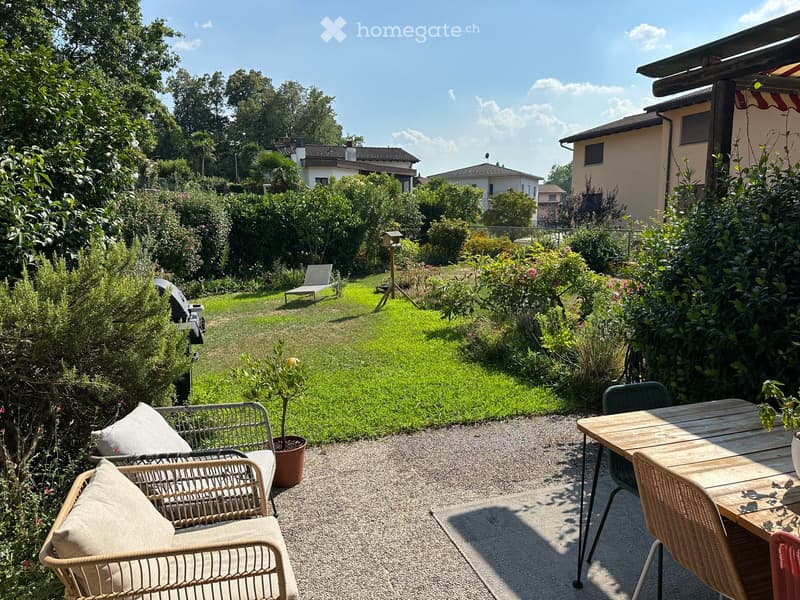 Well-maintained garden with a gravel patio area featuring a wooden table, wicker chairs, and a lounge chair. The garden includes lush greenery, a grassy lawn, and a small potted plant.