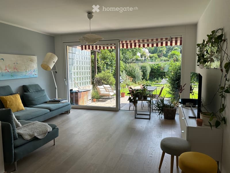 Modern living room with light wood flooring, grey sofa, two yellow stools, a small table, and a hanging plant. Large sliding glass doors open to a patio with outdoor furniture.