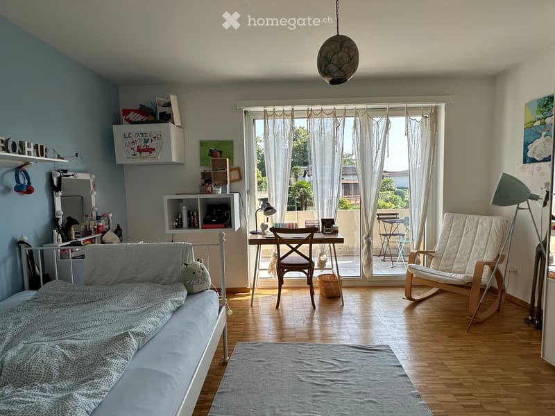A cozy, well-lit room with a sofa, a small table, a rocking chair, a mirror, shelves, and a view of a balcony area.