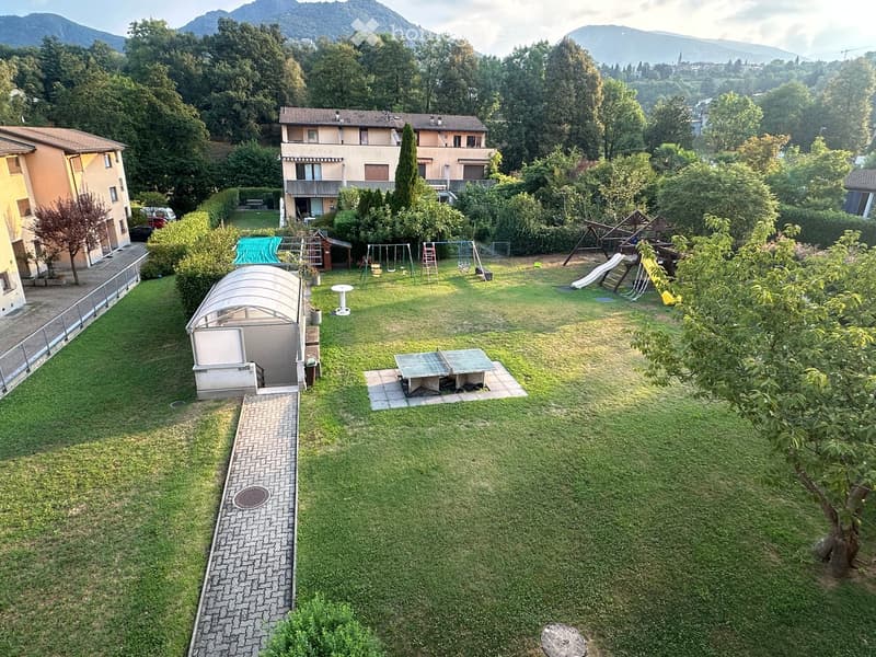 A well-maintained garden with a lawn, a covered patio area, a ping pong table, a playground with swings and a slide, and surrounding trees and shrubs.