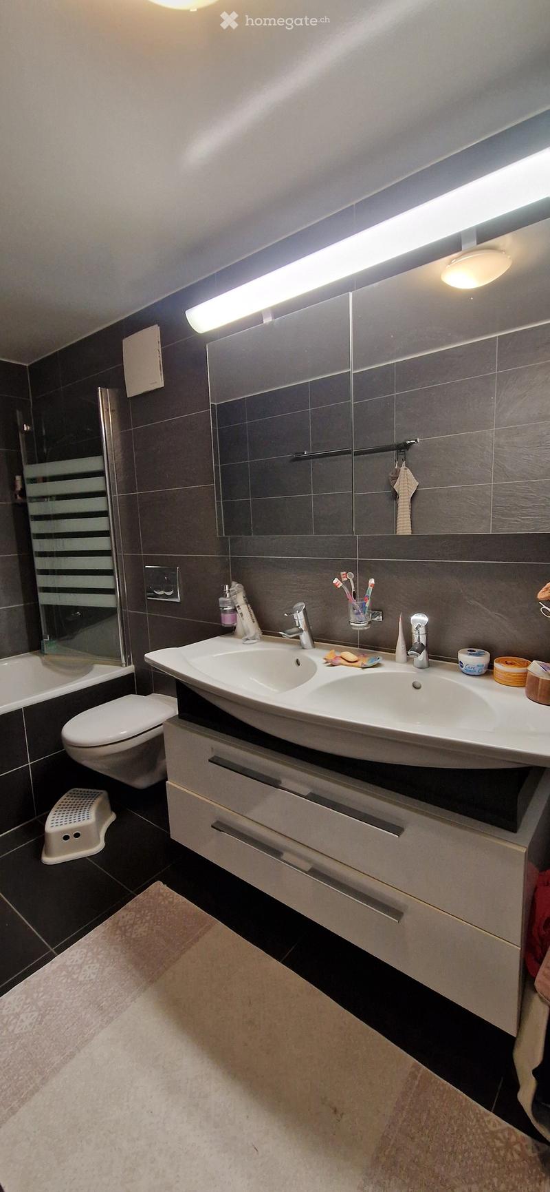 Modern bathroom with dark grey tiles, double sink, mirror, toilet, and a small white stool near the bathtub.
