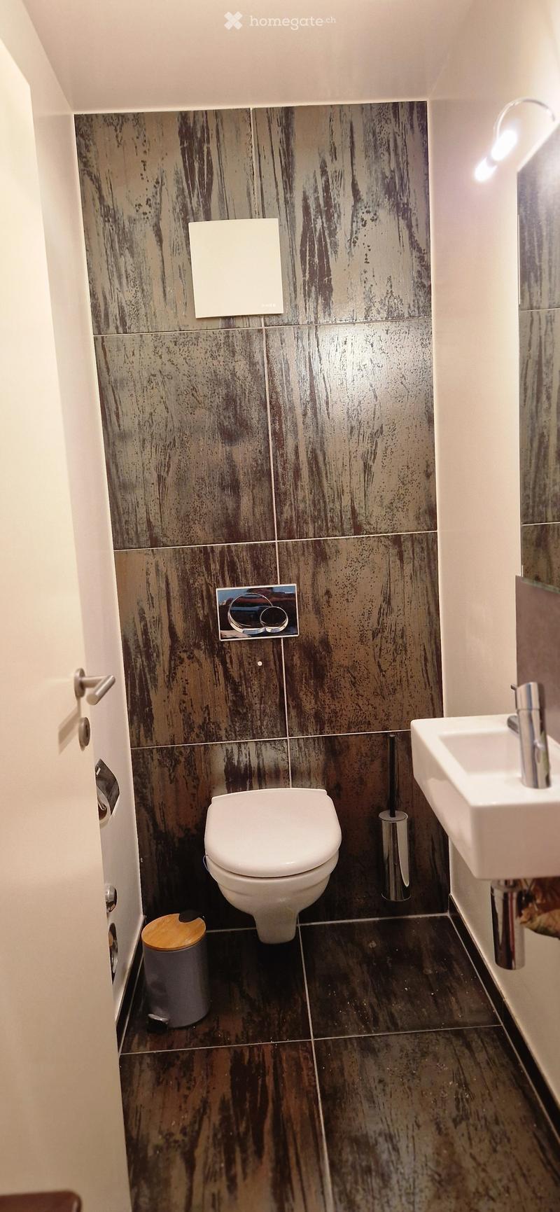 Modern bathroom with dark, textured tiles, a white toilet, a white sink with a chrome faucet, and a small trash can with a wooden lid.