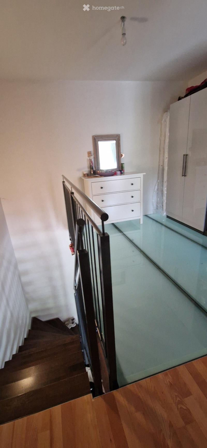 Staircase with wooden steps and a glass railing, leading to a small landing area with a dresser, mirror, and a few decorative items.