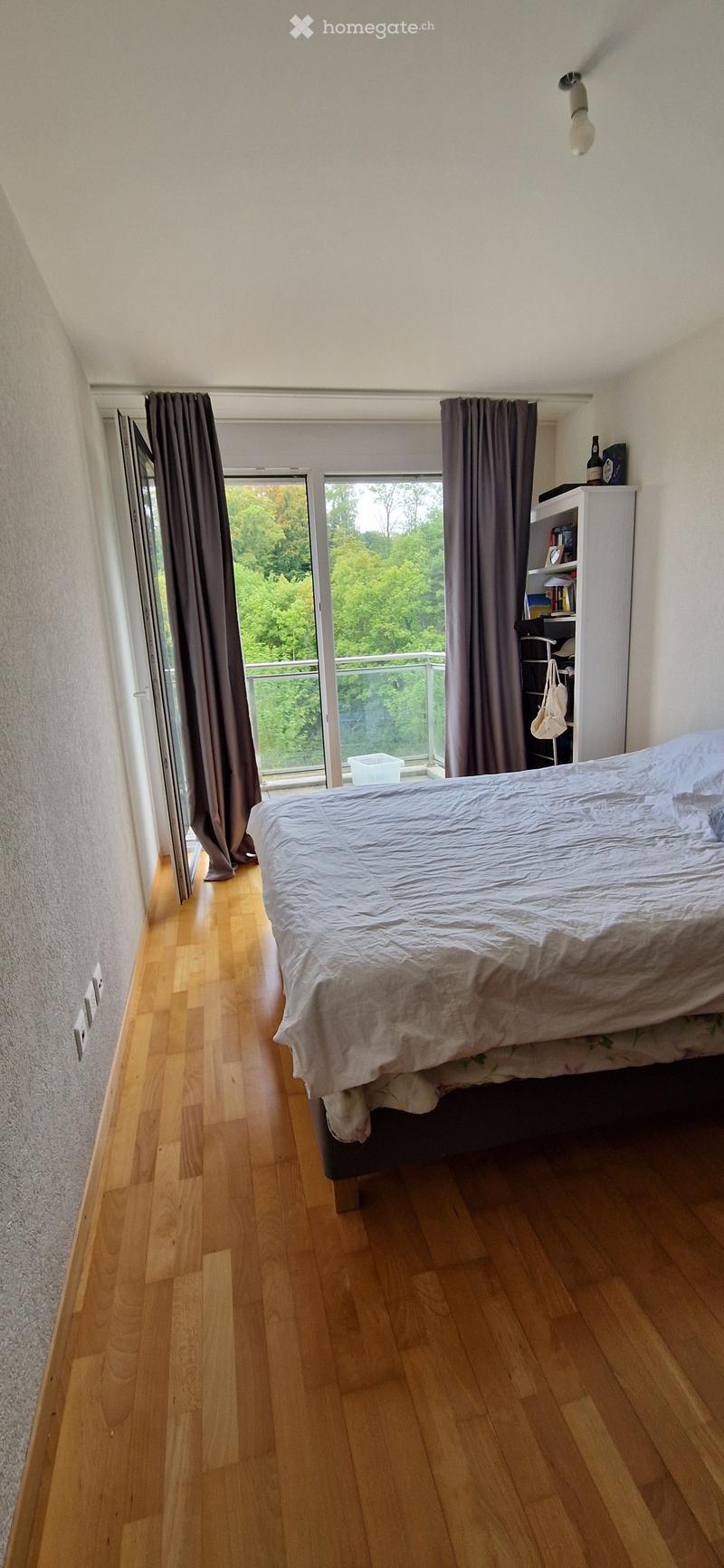 A modern bedroom with wooden flooring, a large bed with white bedding, a bookshelf, and large windows with curtains offering a view of greenery.