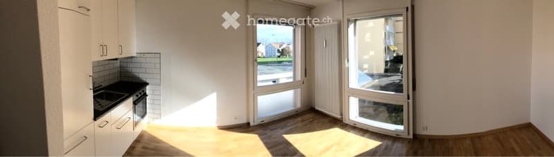 Empty kitchen with white cabinets, a black countertop, and a window overlooking the street. The floor is wooden, and the space is well-lit with natural light.
