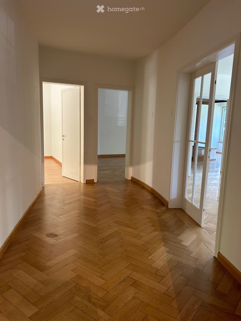 Empty hallway with wooden herringbone flooring, white walls, and multiple doors leading to other rooms.