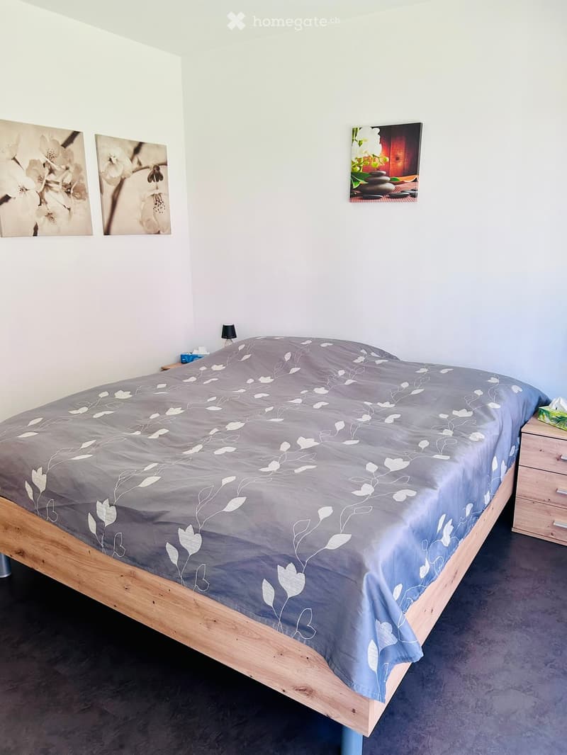 A modern bedroom with a wooden bed frame, grey duvet with leaf pattern, nightstand with lamp, and decorative picture frames on the wall.