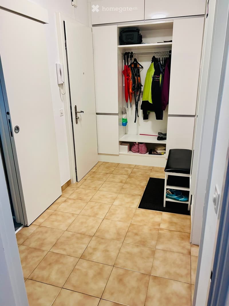 Clean hallway with tiled floor, white walls, built-in closet with clothes and shoes, small bench, and a black doormat.