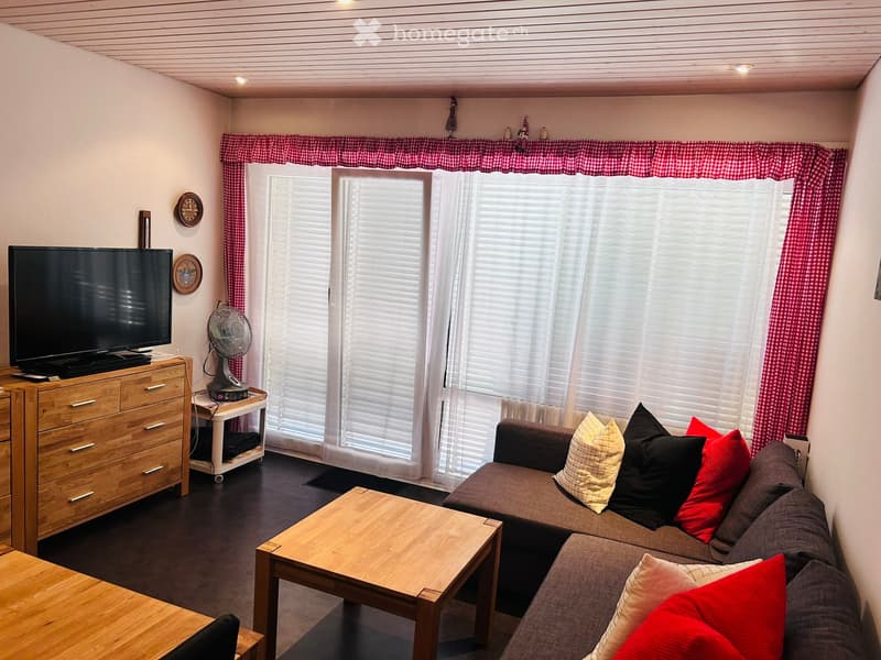 Living room with a sofa, coffee table, television, fan, and a wooden drawer unit. Large window with blinds and red checkered curtains.