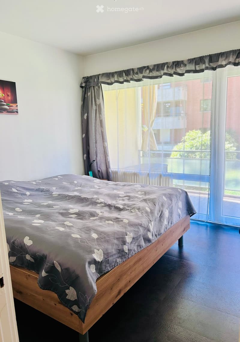 Modern bedroom with a wooden bed, dark grey bedding, and large window with curtains leading to a balcony.