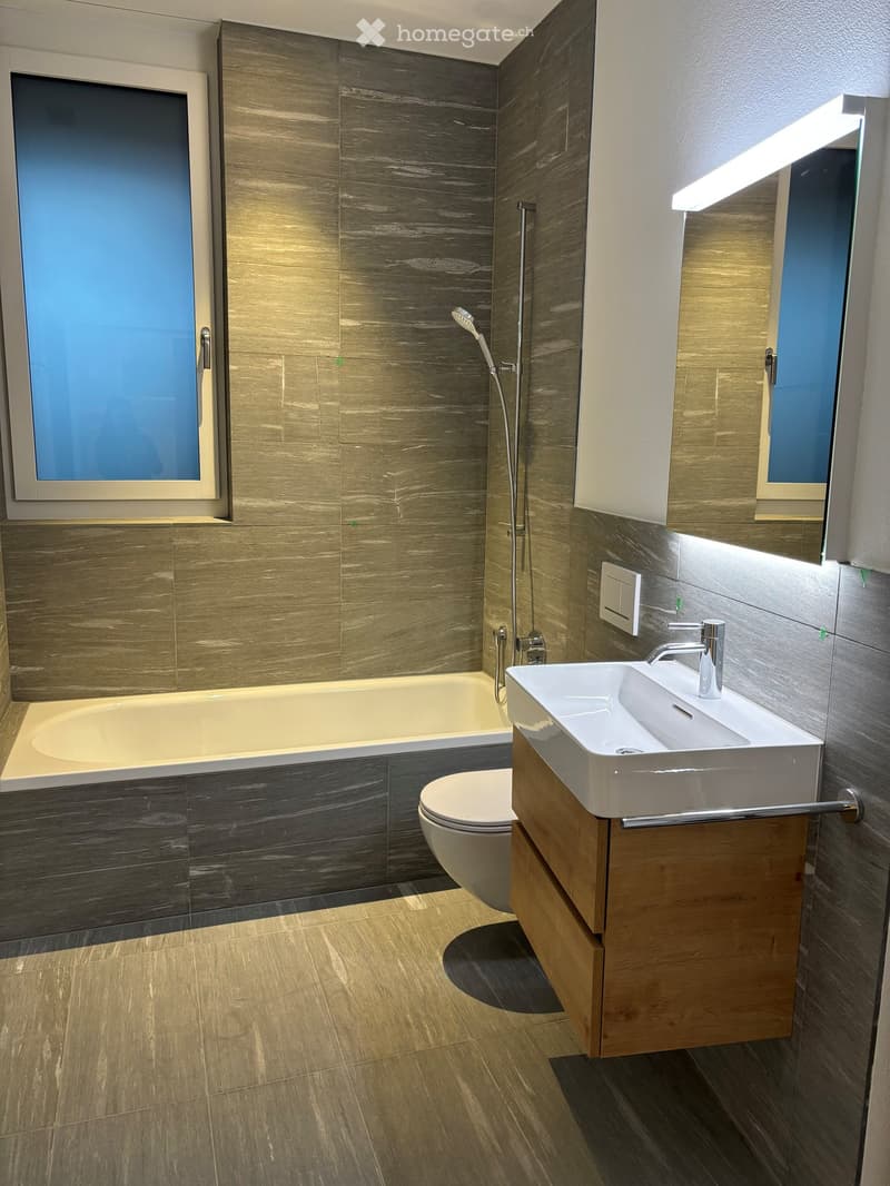 Modern bathroom with a bathtub, toilet, sink, and showerhead. Features wooden vanity, large mirror, and sleek fixtures.