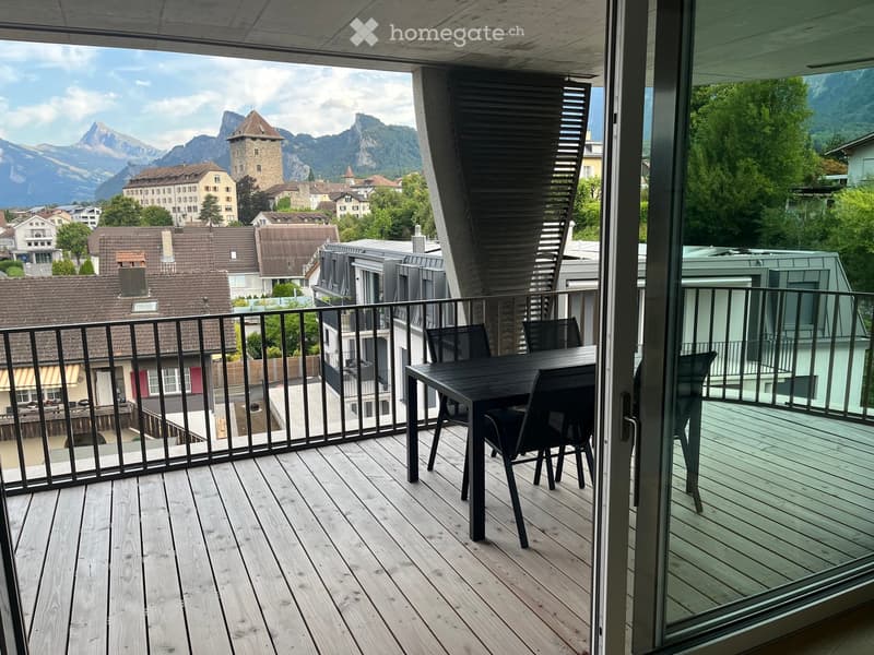 Balcony with wooden decking, black metal railing, and a table with chairs, overlooking a town with mountains in the background.