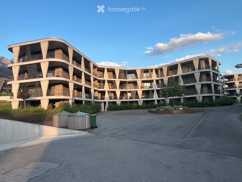 Modern, multi-story residential building with curved balconies and large windows, surrounded by greenery and a paved area.