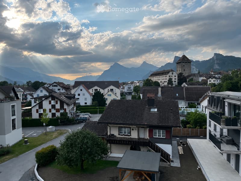 A picturesque town with traditional houses, a prominent tower, and surrounding mountains under a partly cloudy sky.
