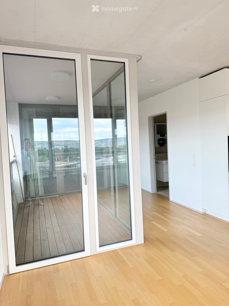 Empty room with light wooden flooring, white walls, and large glass sliding doors leading to a balcony with a view of the city.