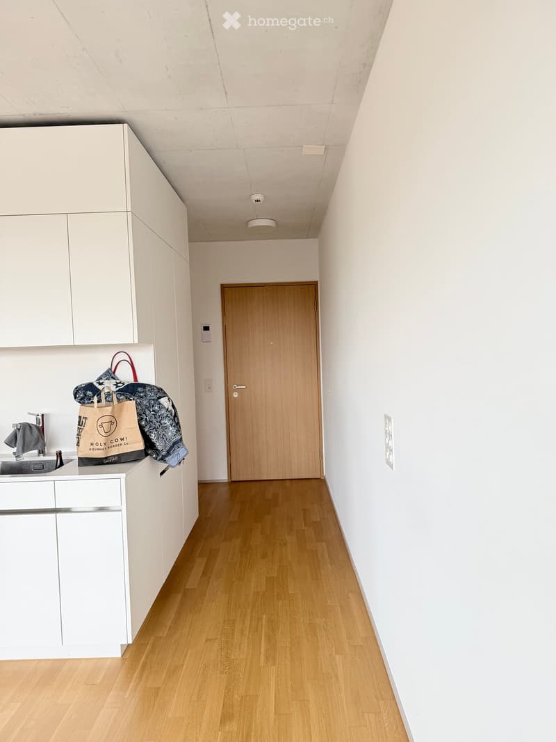 A small, modern kitchen area with white cabinets, a wooden door, and a sink with a faucet. A handbag and a towel are placed on the counter.