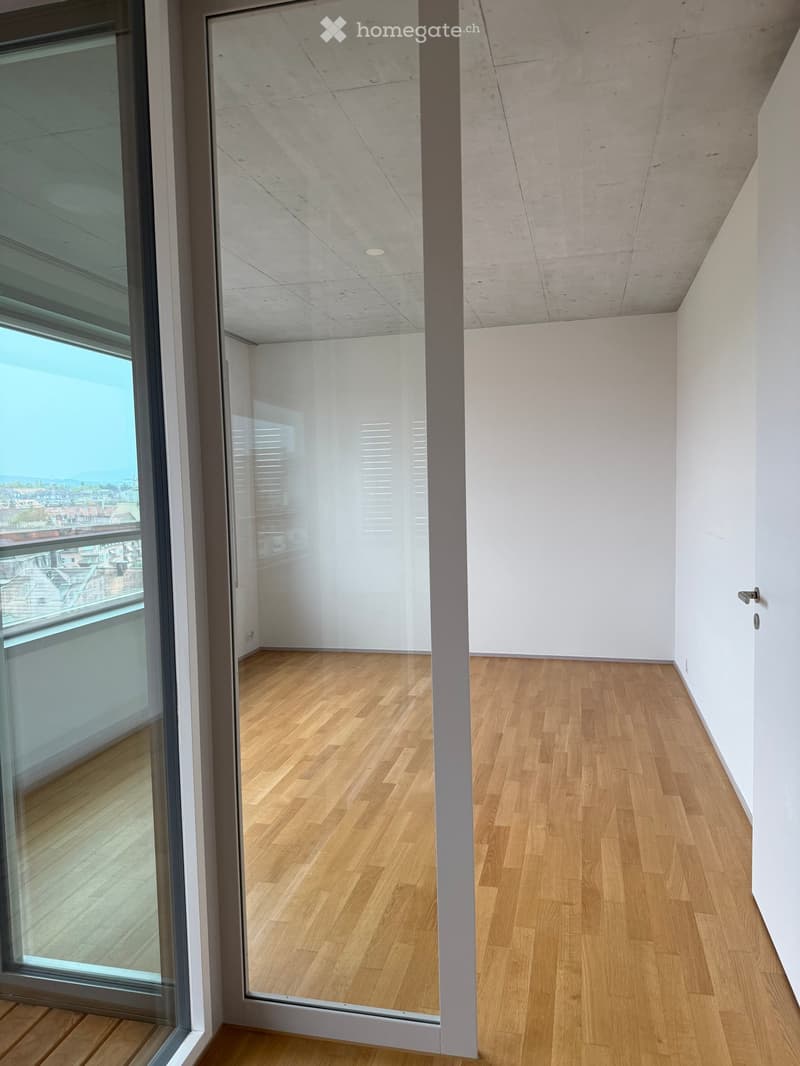 Empty room with light wooden flooring, white walls, and large windows offering a view of the cityscape.