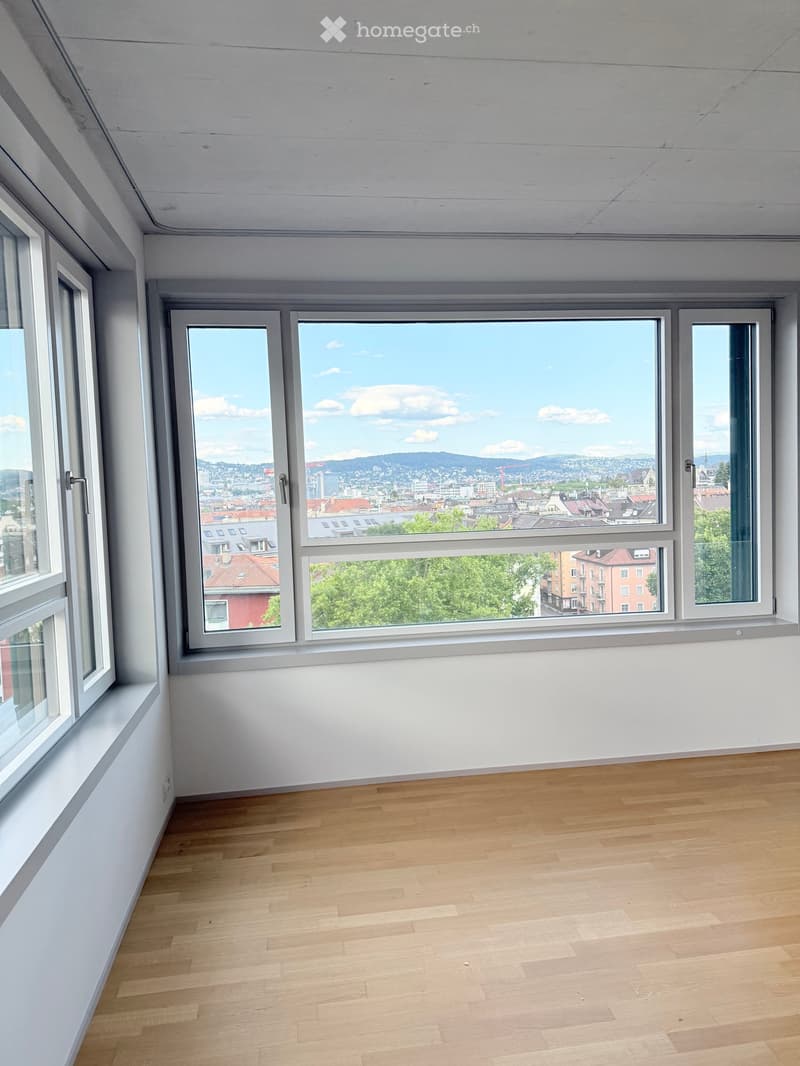 Empty room with light wooden flooring and large windows offering a cityscape view.