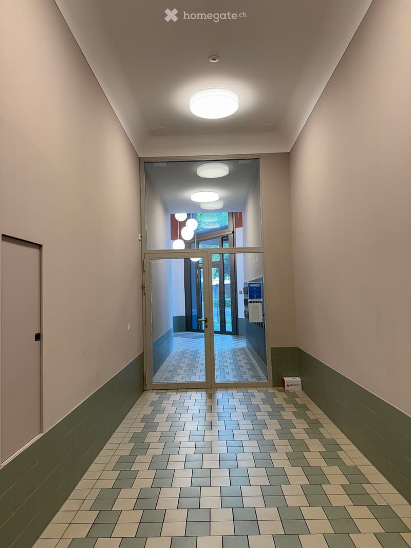 A modern hallway with tiled flooring, light-colored walls, and a large glass door leading to an outdoor area. The ceiling features recessed lighting and a central circular light fixture.