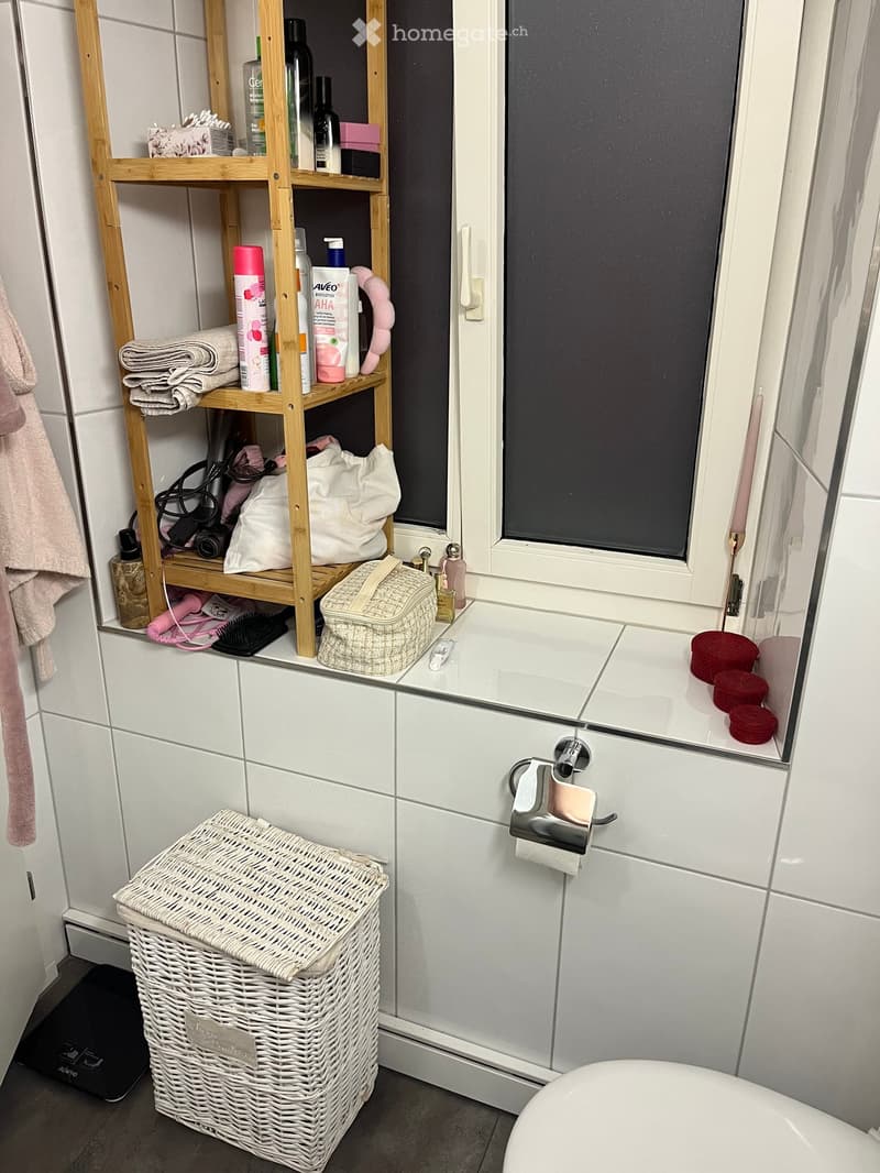 Modern bathroom with white tiles, a wooden shelf holding towels and cosmetics, a wicker basket, and a toilet paper holder.
