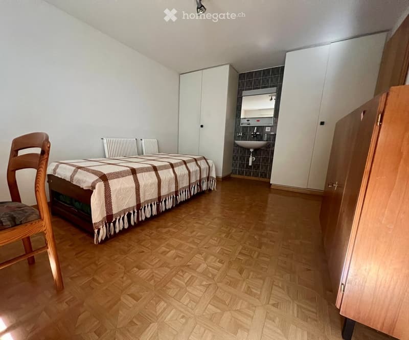 A small, furnished room with a bed, a chair, a sink, and a wardrobe. The room features wooden flooring and white walls.