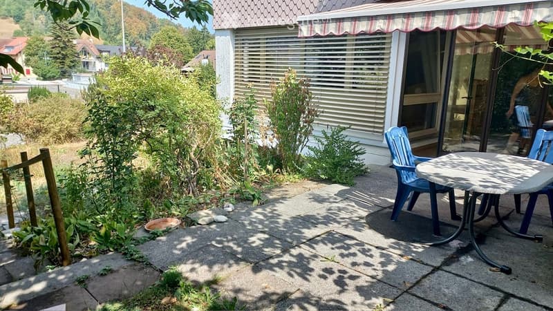 Patio area with a round table and two blue chairs, surrounded by greenery and a house with a striped awning.