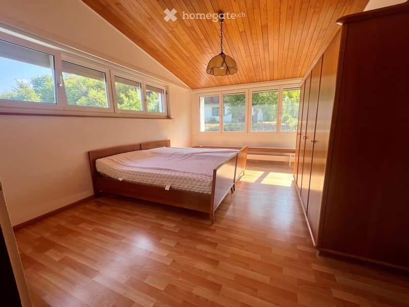 Bright bedroom with wooden floors, slanted wooden ceiling, large windows, bed with white bedding, wardrobe, and bench.