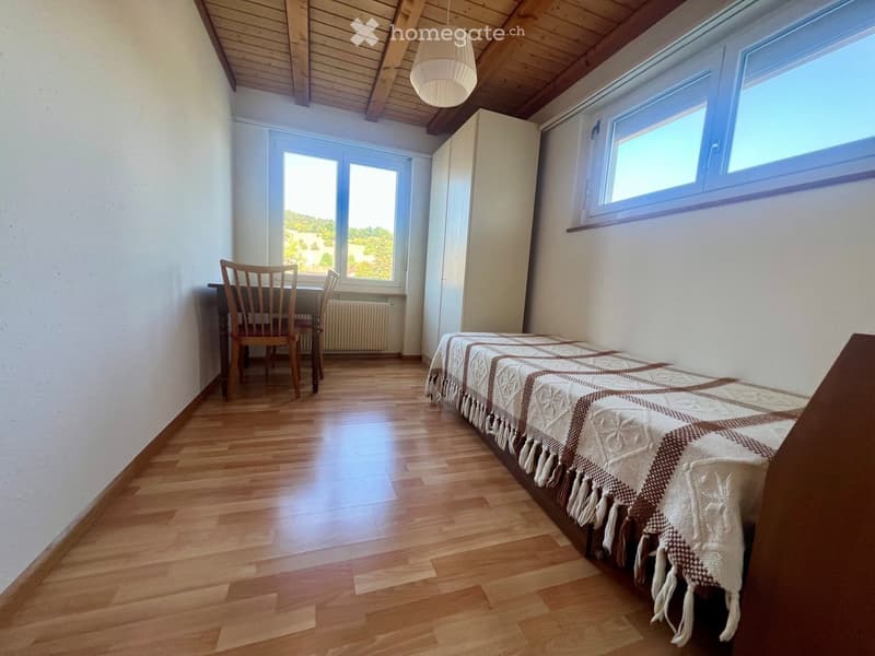 Cozy bedroom with wooden flooring, a bed with a patterned blanket, a small table with two chairs, and large windows offering a scenic view.