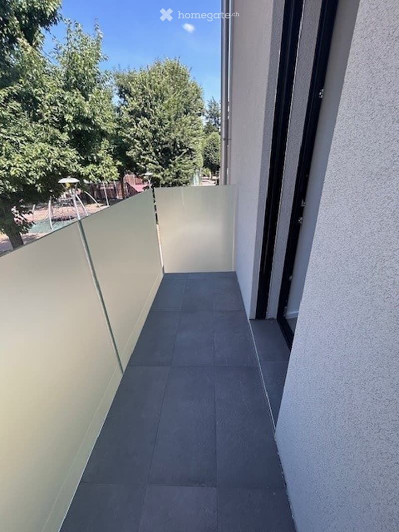 Modern balcony with grey tiled flooring, white walls, and glass balustrade. The balcony is enclosed and offers a view of a green area with trees.