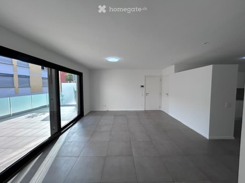 Completely empty room with bare walls, tiled floor, large window, and white door.