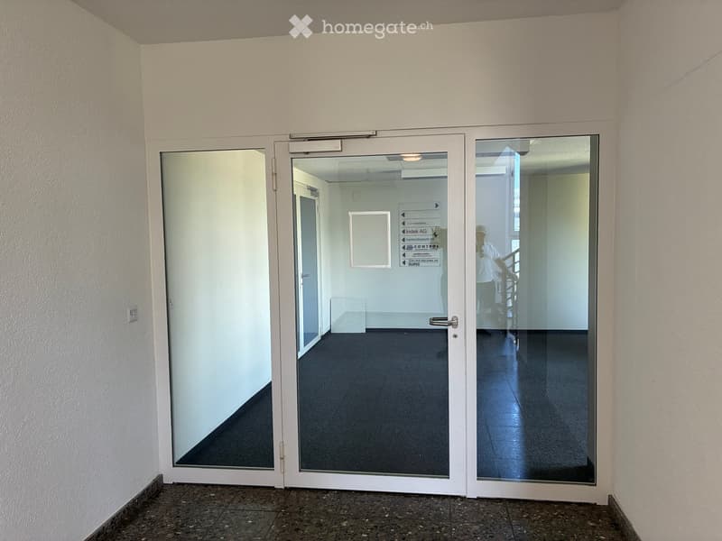 A clean, modern hallway with white walls, dark tiled flooring, and glass doors leading to an open space. The glass doors have directional signs and a visible reflection of a person in the background.