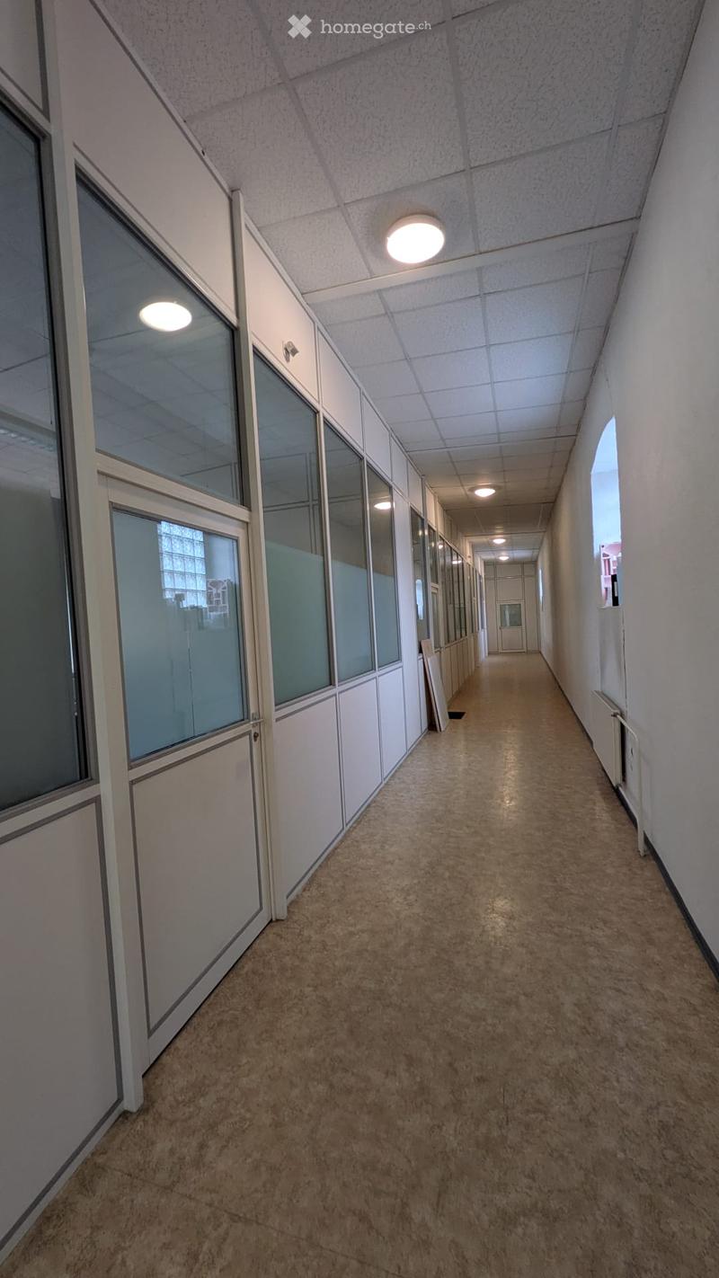 A long, clean hallway with beige tiled flooring, white walls, and recessed lighting. Glass partitions line one side, and a radiator is visible on the right wall.