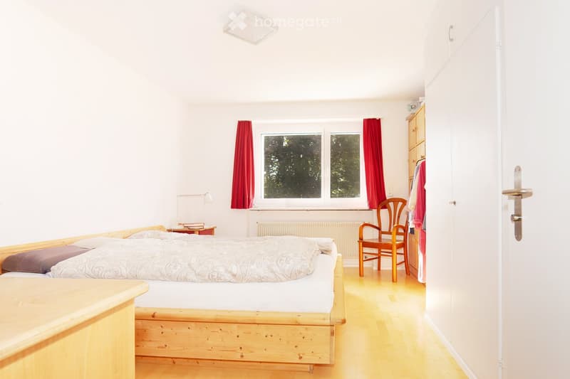 a wooden bed with white sheets, red window curtains, wooden chair, radiator, white walls, and wooden floor