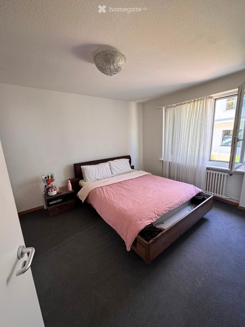 A well-lit bedroom with a wooden bed, pink bedding, a nightstand with toiletries, a wicker hanging lamp, and a window with white curtains.