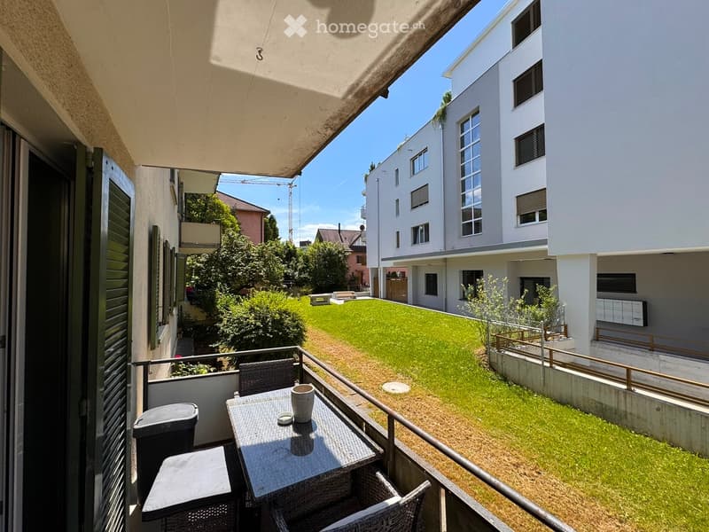 Balcony with a small table, chairs, and a cup, overlooking a green lawn and modern buildings.