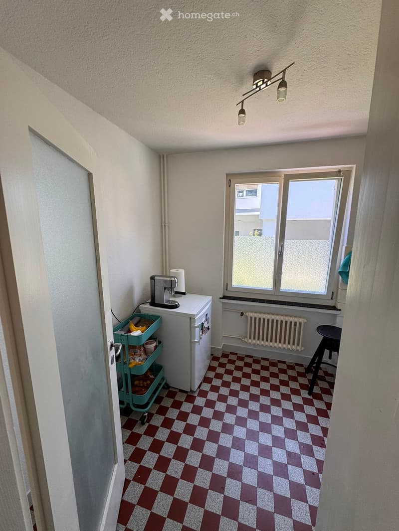 Small kitchen with checkered tile floor, white refrigerator, coffee maker, green cart, and a stool near the window.